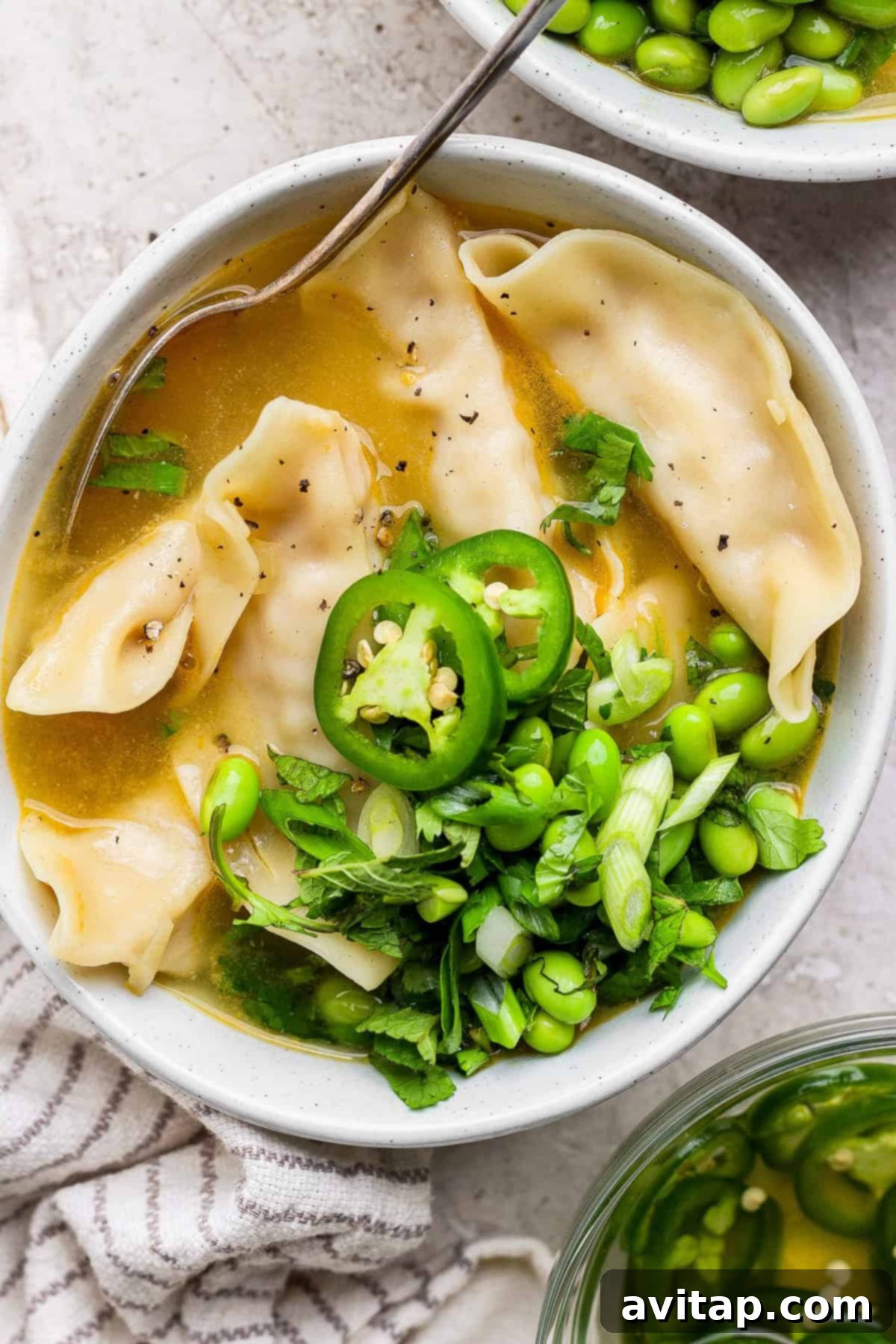 Zesty Wonton Delight 2 A bowl of broth with dumplings, sliced green chili, edamame, and chopped green onions, garnished with herbs. A spoon is resting in the bowl.