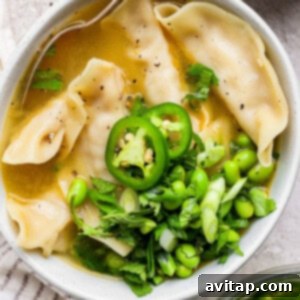 Zesty Wonton Delight 11 A bowl of broth with dumplings, sliced green chili, edamame, and chopped green onions, garnished with herbs. A spoon is resting in the bowl.
