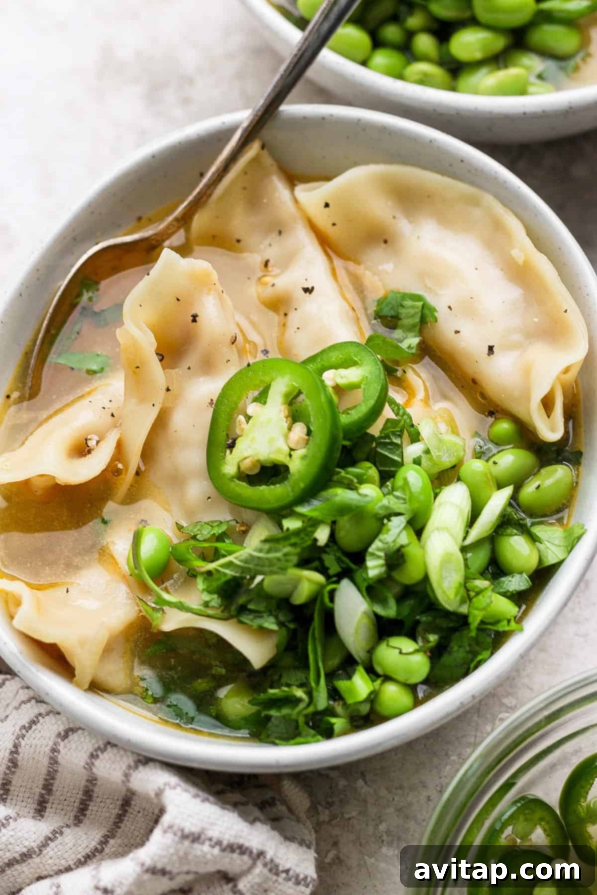 Zesty Wonton Delight 12 A bowl of soup with dumplings, sliced jalapeños, green onions, edamame, and cilantro. A spoon rests in the bowl, and a striped cloth is nearby.
