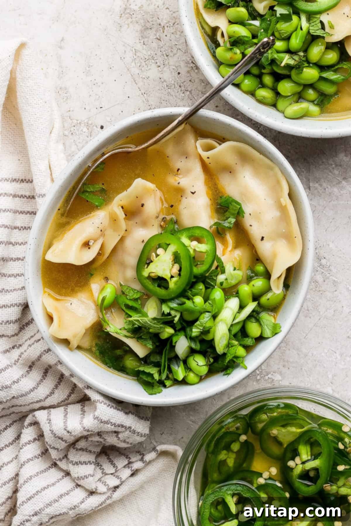 Zesty Wonton Delight 13 A bowl of dumpling soup garnished with sliced jalapeños, chopped green onions, and cilantro, with a side of edamame. A striped cloth is beside the bowl.