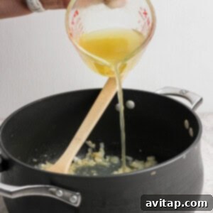 Zesty Wonton Delight 7 A person pours yellow liquid from a measuring cup into a black pot containing sautéed onions, with a wooden spoon resting in the pot.