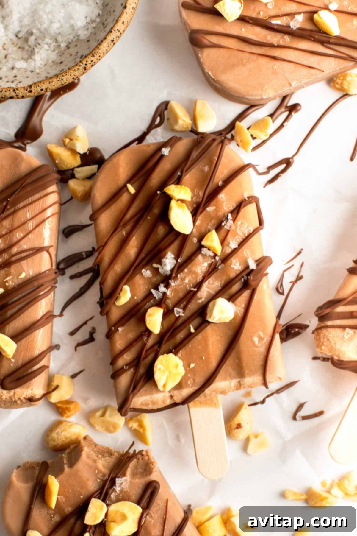 Chocolate popsicles drizzled with chocolate and sprinkled with chopped nuts and sea salt on a white surface.