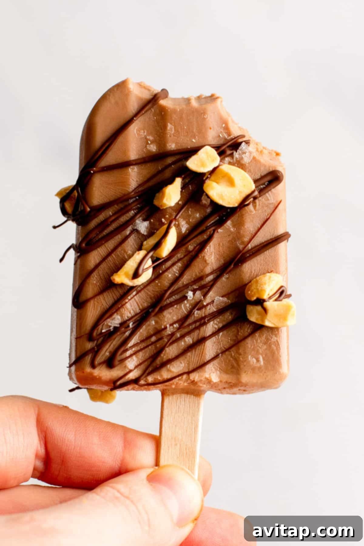 Person holding a chocolate popsicle with drizzled chocolate and peanut pieces, partially eaten.