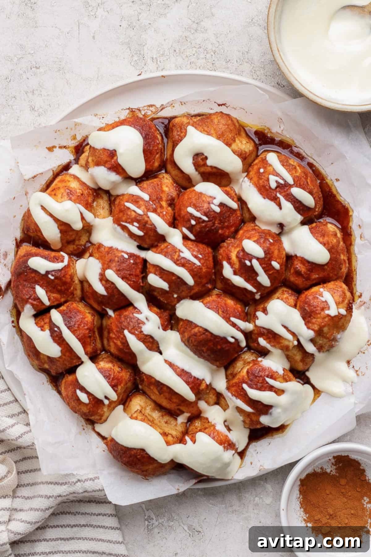 A round cinnamon pull-apart bread topped with cream cheese icing on parchment paper. Nearby are a bowl of icing and a dish of cinnamon.