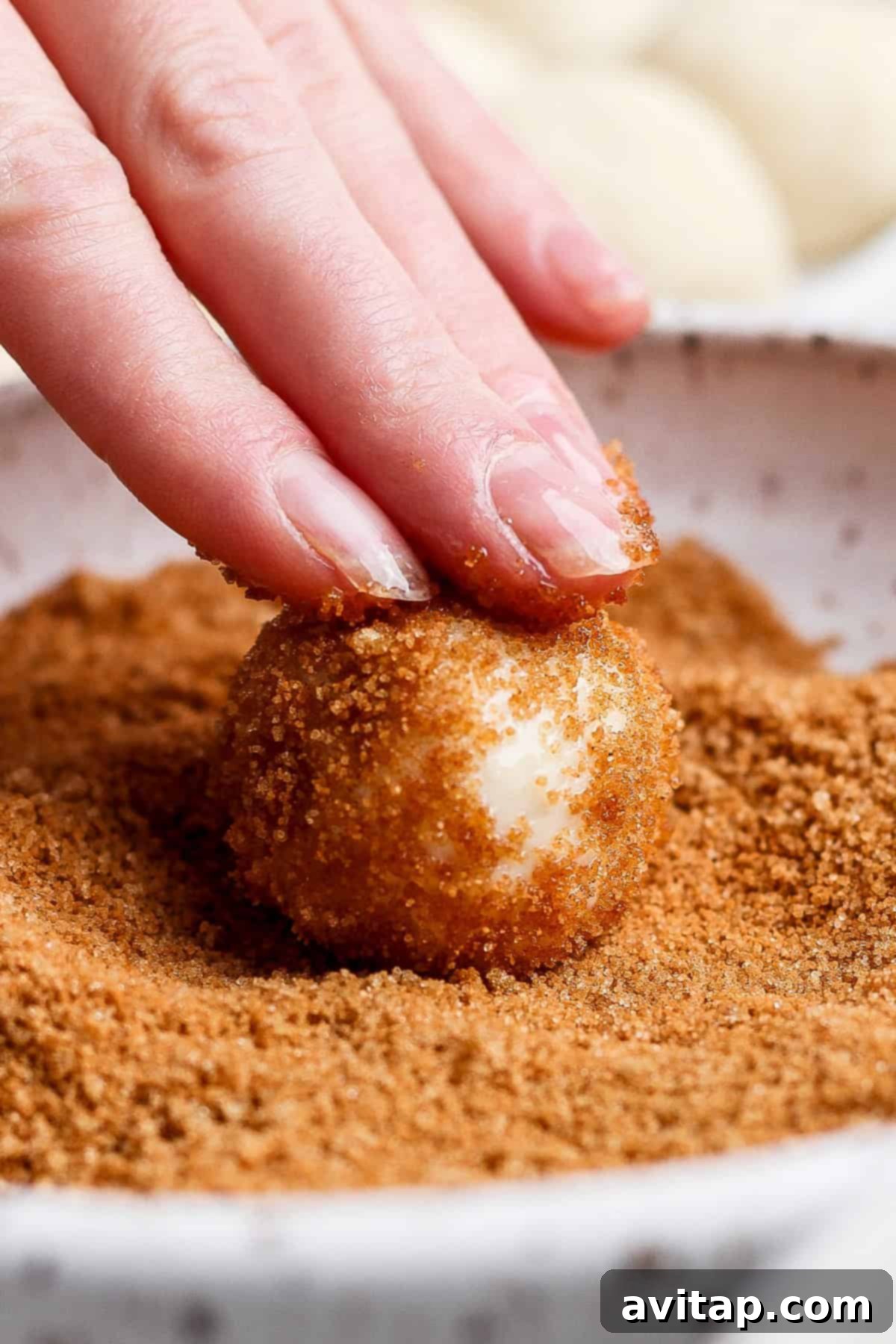 A hand rolls a dough ball in a bowl of brown sugar topping.