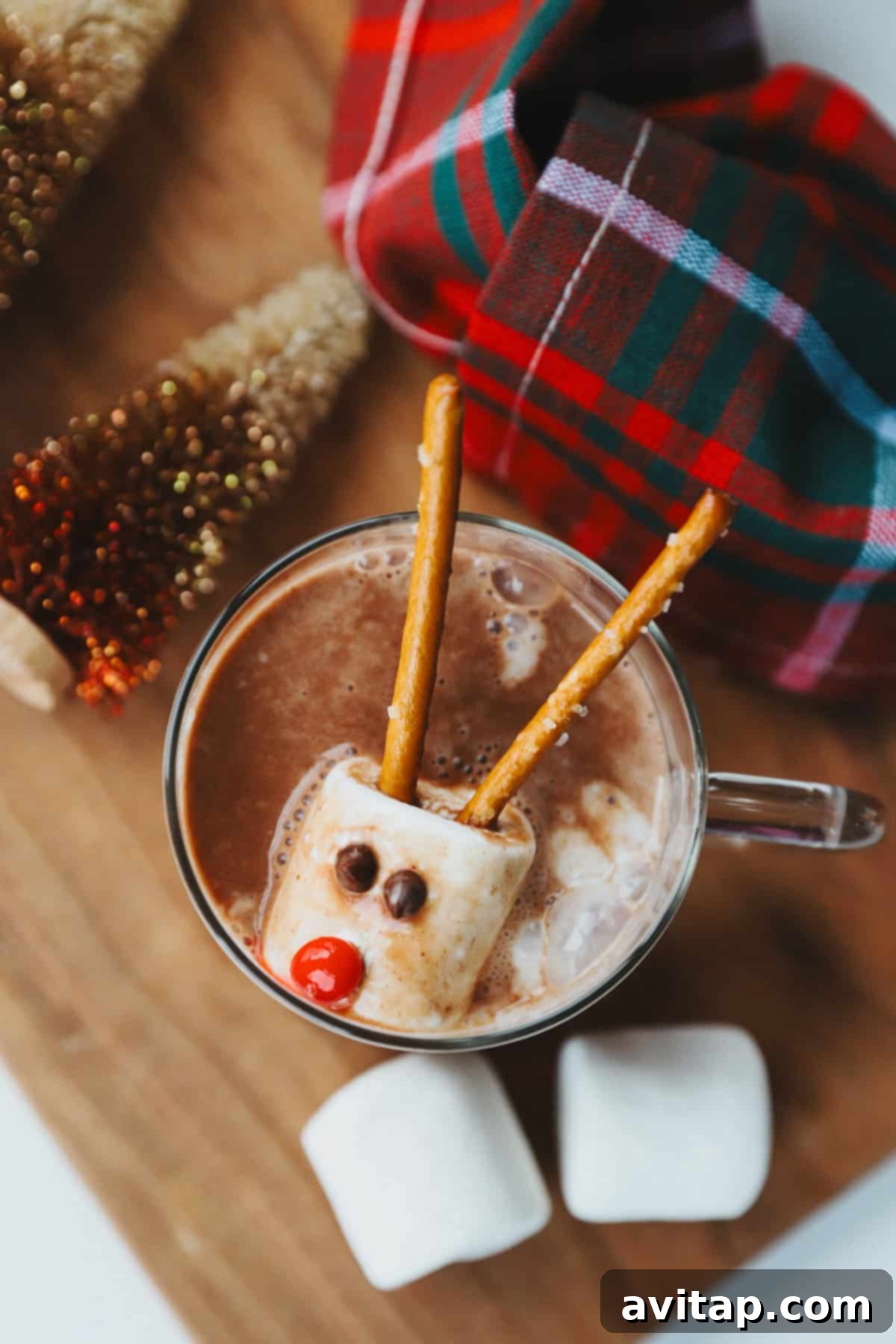 Whimsical Snowman Melts 2 A charming cup of hot chocolate, garnished with a marshmallow reindeer featuring pretzel antlers, candy eyes, and a festive red candy nose, positioned beside a cozy plaid fabric and miniature Christmas trees. This image perfectly captures the spirit of holiday treats.