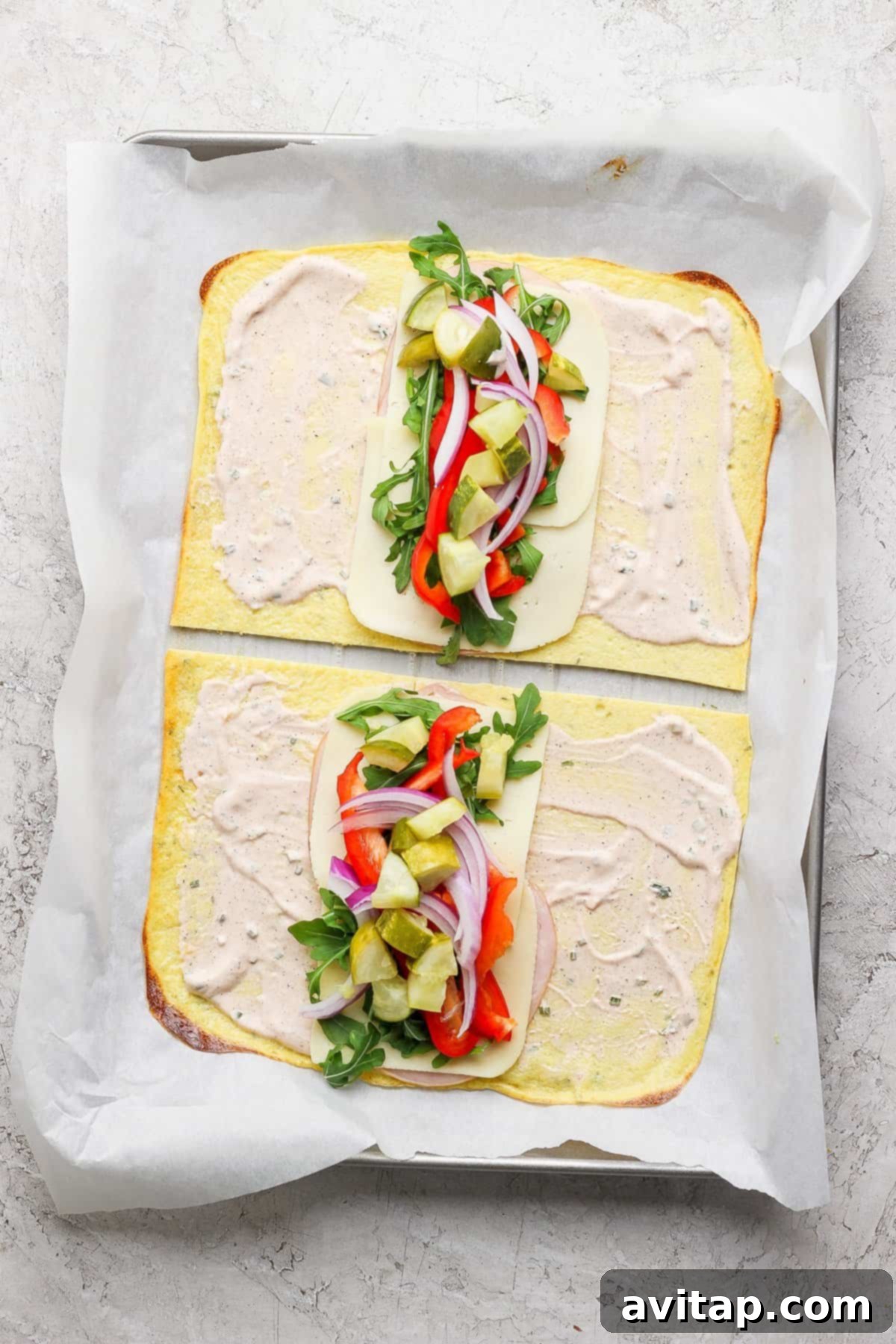 Two open-faced flatbread sandwiches on a baking sheet, with cheese, arugula, red onions, red peppers, and pickles, topped with a creamy spread, placed on parchment paper.