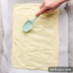Hand spreading batter evenly on a baking sheet lined with parchment paper using a teal spatula.