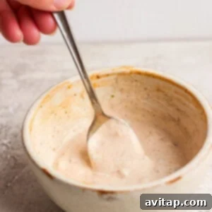 Hand holding a spoon in a ceramic bowl of creamy sauce on a gray surface.