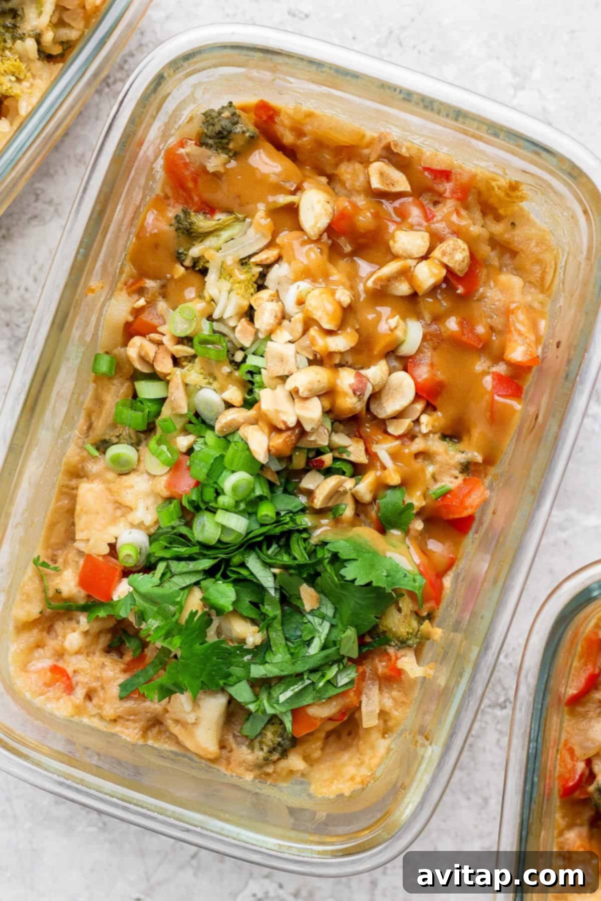 A glass container holds a colorful salad topped with chopped peanuts, green onions, and cilantro, accompanied by tender slices of peanut chicken, resting elegantly on a light textured surface.