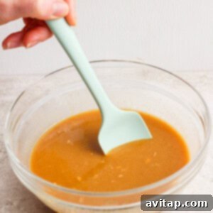 A hand stirs brown gravy in a clear glass bowl with a light green spatula on a light surface.