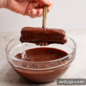 A hand holds chopsticks dipping a chocolate-covered rectangular treat into a glass bowl of melted chocolate.
