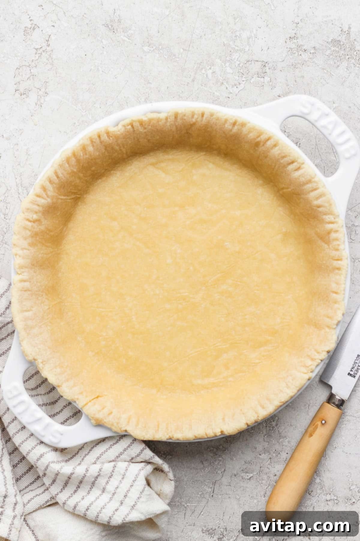 Wholesome Protein Pie Crust 2 Unbaked high-protein pie crust in a pristine white ceramic pie dish, resting on a rustic textured surface. A sharp knife and a striped kitchen cloth are artfully placed nearby, hinting at the culinary journey ahead.