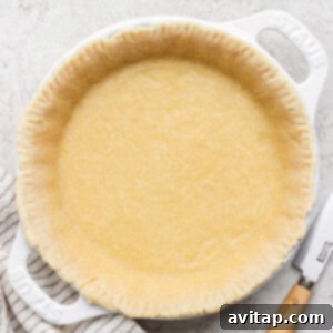 An unbaked protein-rich pie crust nestled in a white ceramic pie dish, accompanied by a striped cloth and a knife on a textured surface, ready for baking.