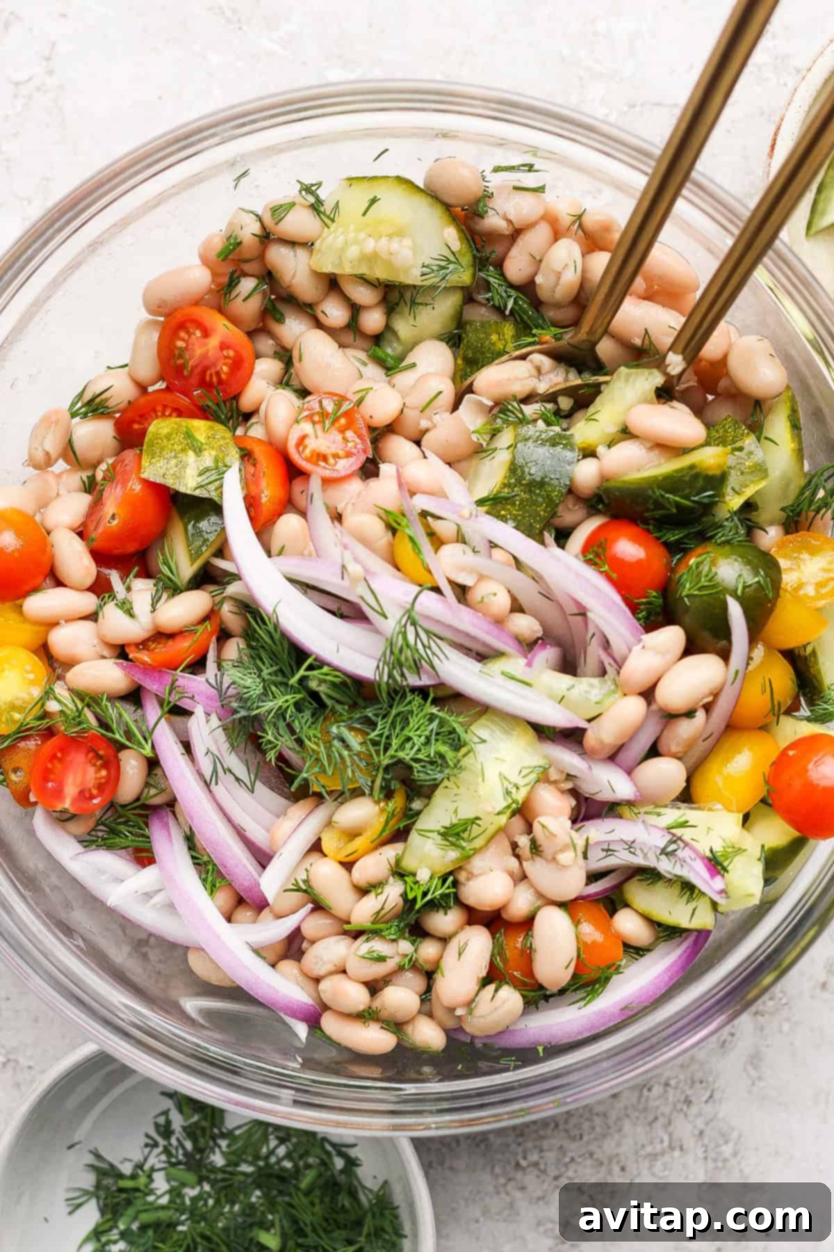 A vibrant bowl of Dill Pickle White Bean Salad featuring white beans, halved cherry tomatoes, thinly sliced red onion, crisp cucumber, and fresh dill, all tossed in a light dressing and garnished with extra herbs. Serving utensils rest in the bowl, inviting you to dig in.