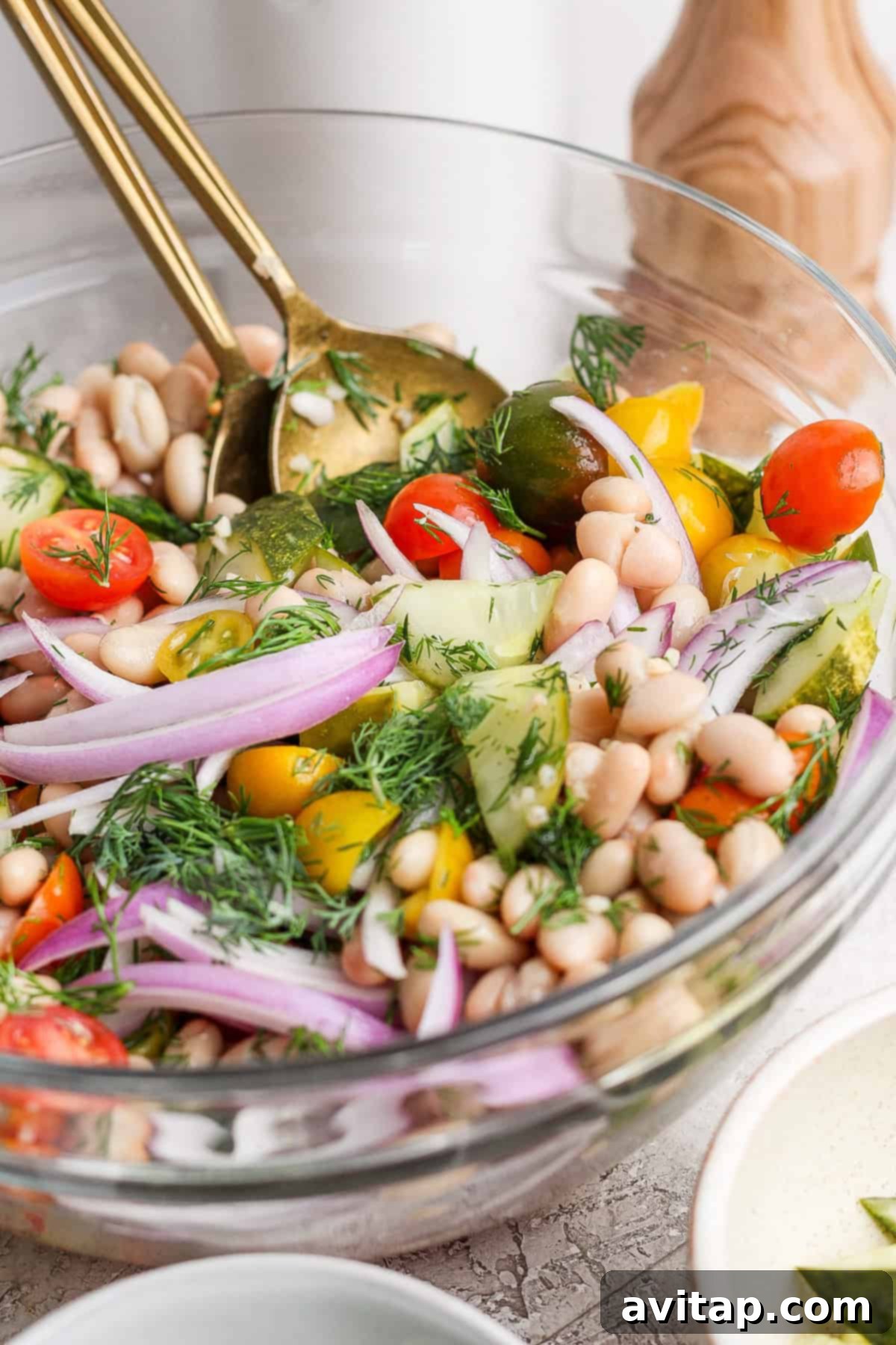 A beautifully presented bowl of vegetarian salad, rich with colorful ingredients including cherry tomatoes, crisp cucumber, creamy white beans, thinly sliced red onion, and fresh dill, artfully tossed with two elegant gold serving utensils.
