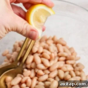 A hand squeezing a lemon wedge over a bowl of white beans, with a gold spoon resting inside the bowl.