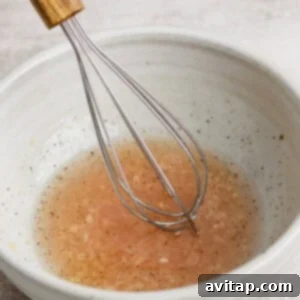 A metal whisk rests in a white bowl filled with a light brown liquid, possibly a vinaigrette or sauce, set on a textured surface.