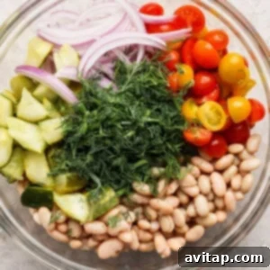 Glass bowl with sliced red onions, halved cherry tomatoes, cucumber chunks, white beans, and a mound of fresh dill.