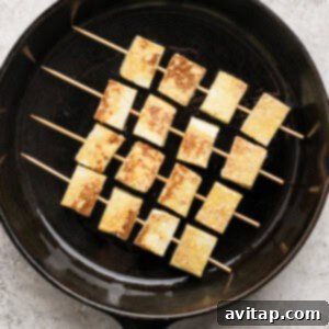 Skewered tofu pieces are grilled in a black cast iron pan on a textured light gray surface.