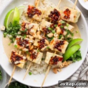 Tofu skewers with rice, garnished with lime wedges, cilantro, chopped nuts, and a drizzle of chili sauce, served in a white bowl.