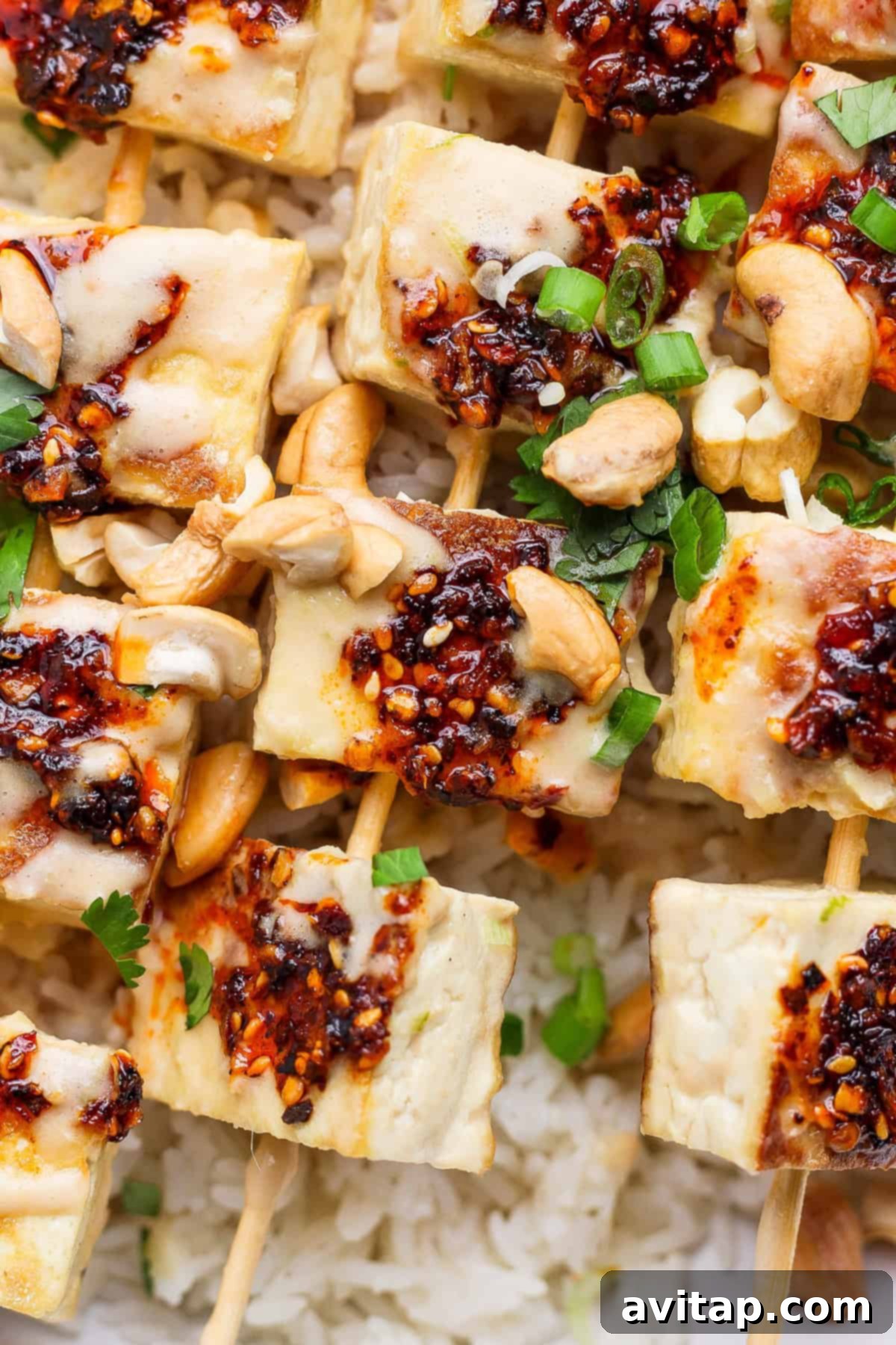 Grilled tofu skewers topped with chili sauce, garnished with green onions and cashews, served on a bed of rice.