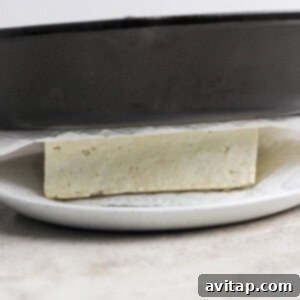 A block of tofu being pressed between a skillet and white plate, with paper towels underneath.