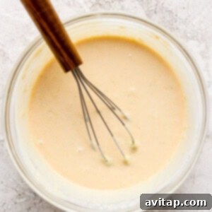 A glass bowl filled with a light beige batter. A whisk with a wooden handle rests in the batter. The surface beneath the bowl is textured and light-colored.