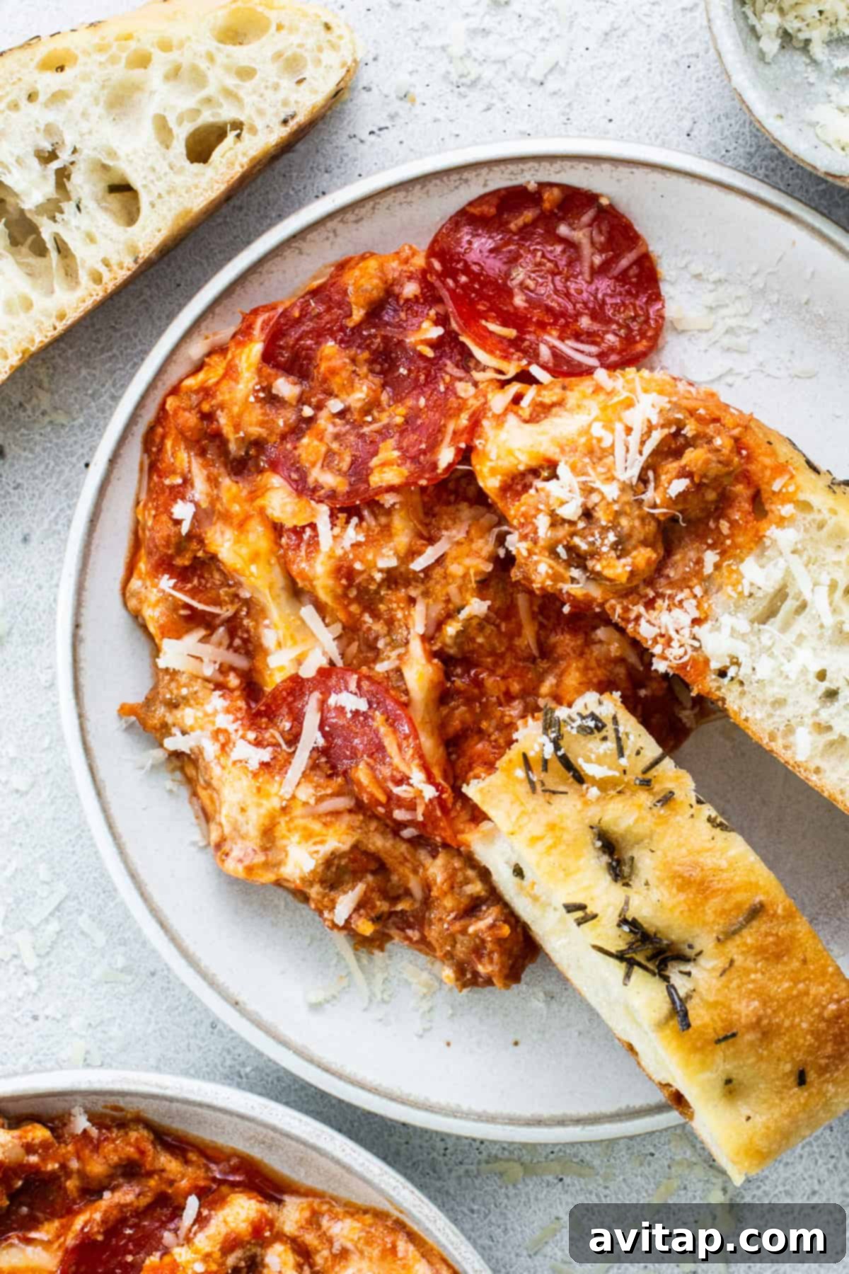 A plate of pepperoni pizza dip topped with cheese, accompanied by slices of crusty bread and rosemary focaccia.