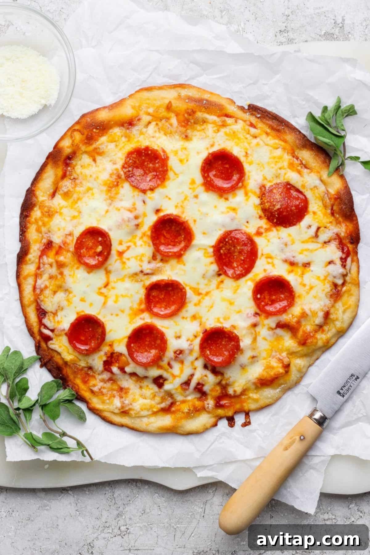 Quick 4 Ingredient Protein Pizza Dough 5 A pepperoni pizza on parchment paper with a knife beside it, garnished with herbs. A small bowl of grated cheese is in the top-left corner.