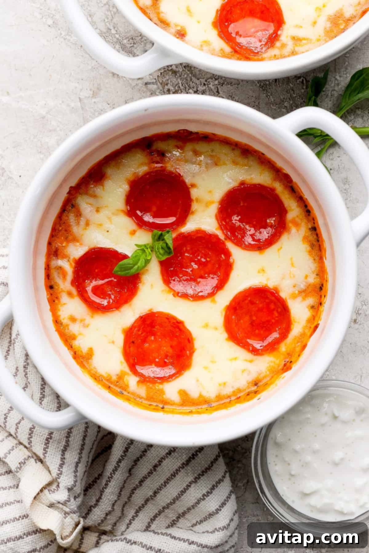 A beautifully baked pepperoni and cheese dip in a white bowl, garnished with fresh basil, placed on a textured cloth next to a small bowl of dipping sauce. This highlights the delicious, melted cheese and crispy pepperoni.