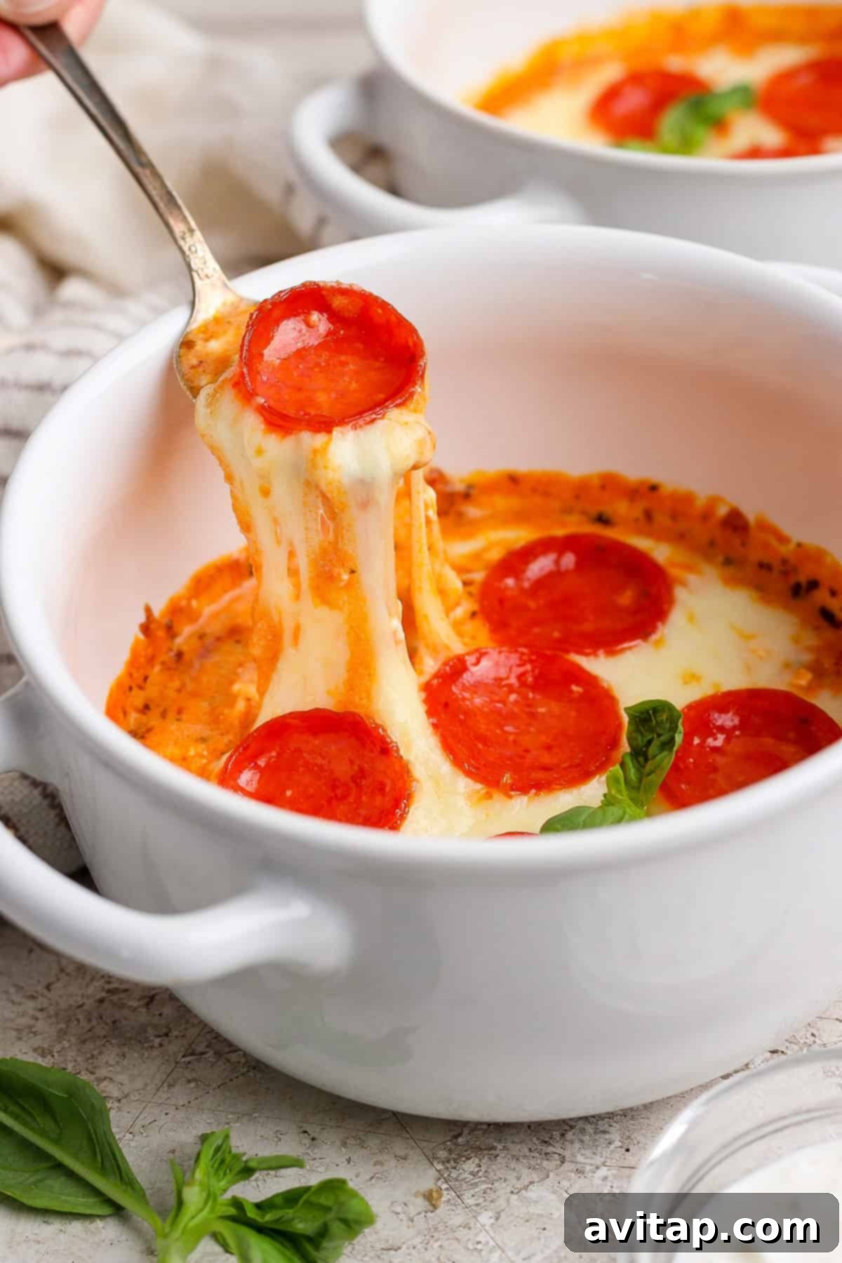 A spoon scooping up a portion of creamy, cheesy pepperoni pizza dip from a white bowl. Fresh basil leaves are visible on the textured surface, suggesting a delicious and aromatic experience.