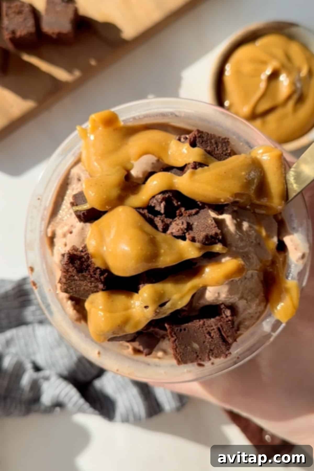 Smooth Chocolate Brownie Protein Dream 2 A cup of chocolate ice cream topped with caramel sauce and brownie chunks, with a spoon in it. A bowl with caramel is visible in the background.