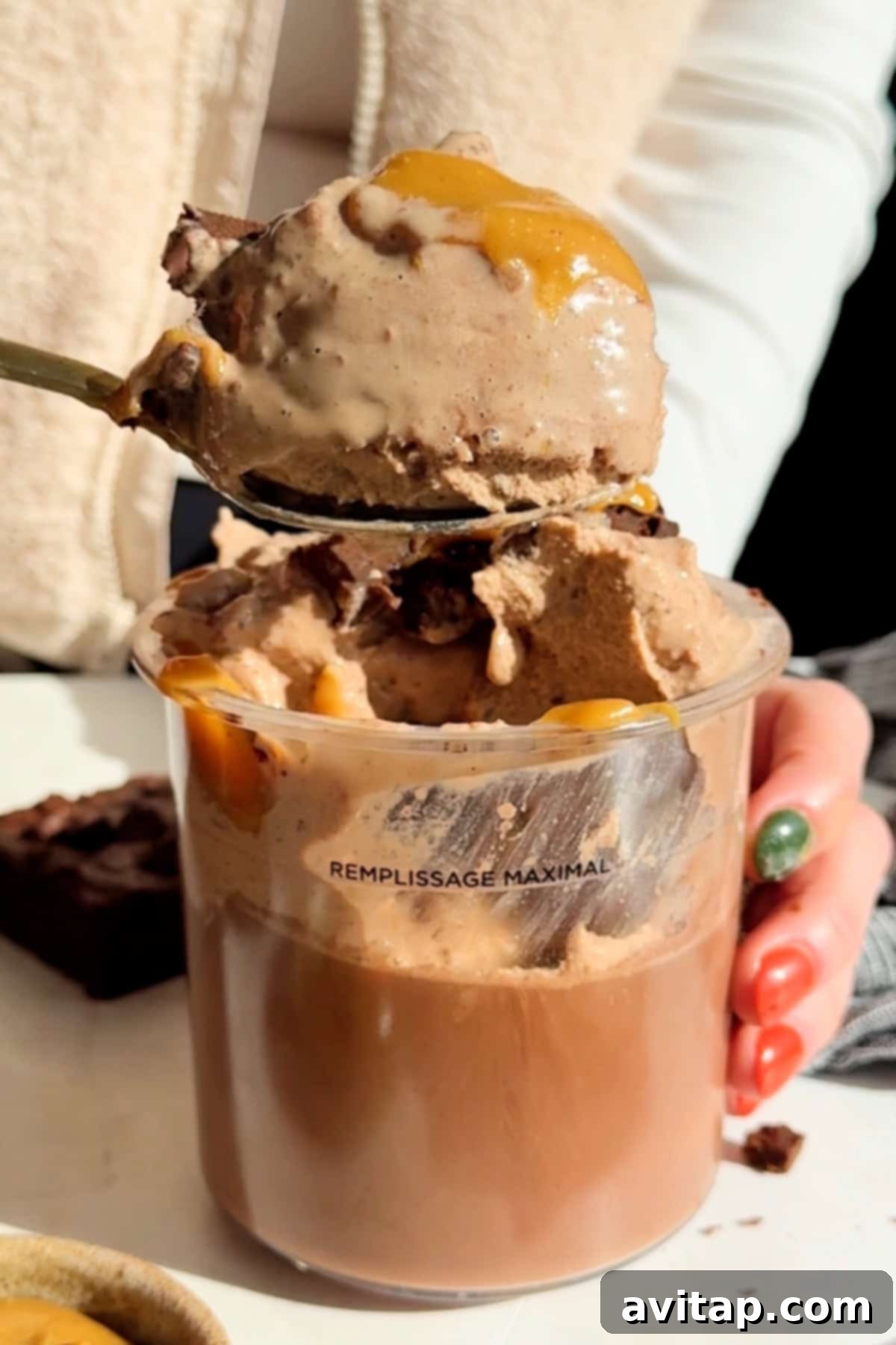 Smooth Chocolate Brownie Protein Dream 4 A person holds a spoon with ice cream drizzled with caramel over a clear cup, which seems filled to the maximum line. A brownie is visible in the background.