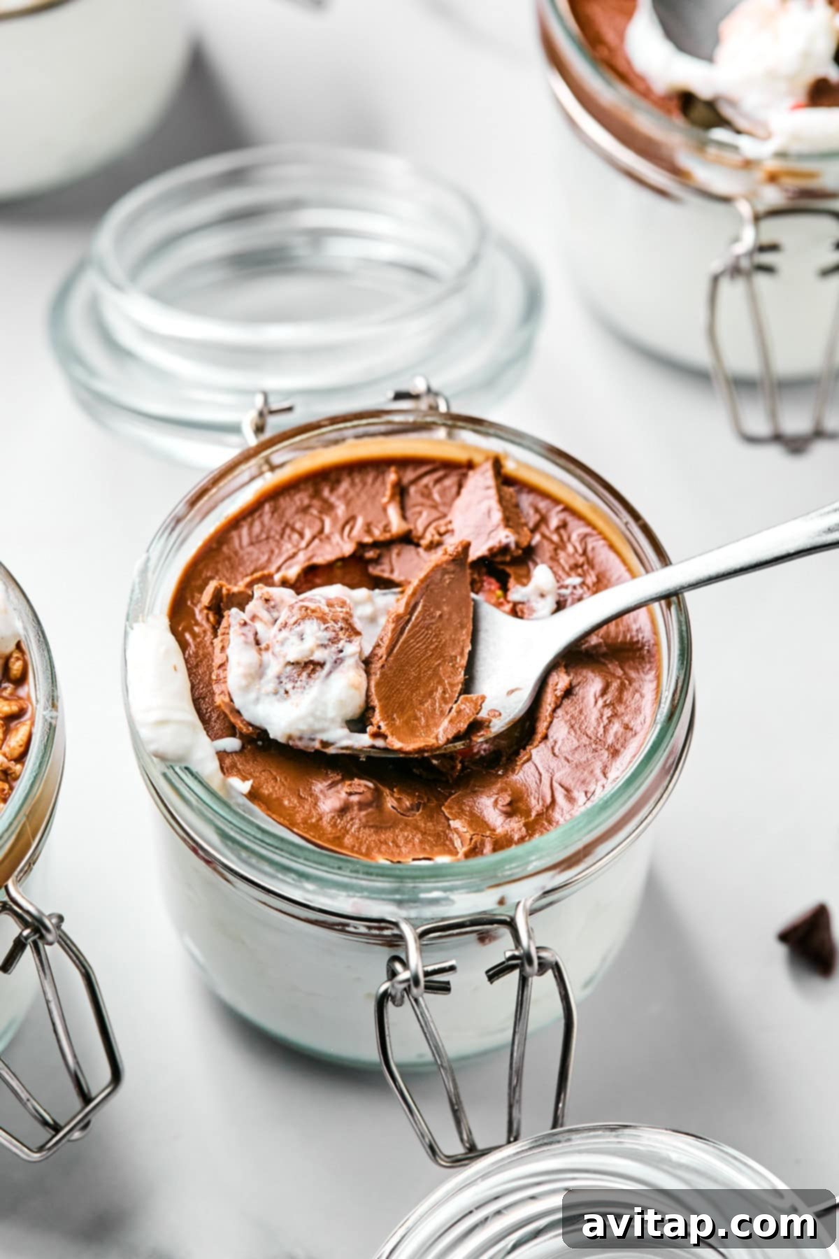 Spoon dipping into a glass jar of chocolate mousse with visible layers of cream and chocolate. Other jars are partially visible in the background.