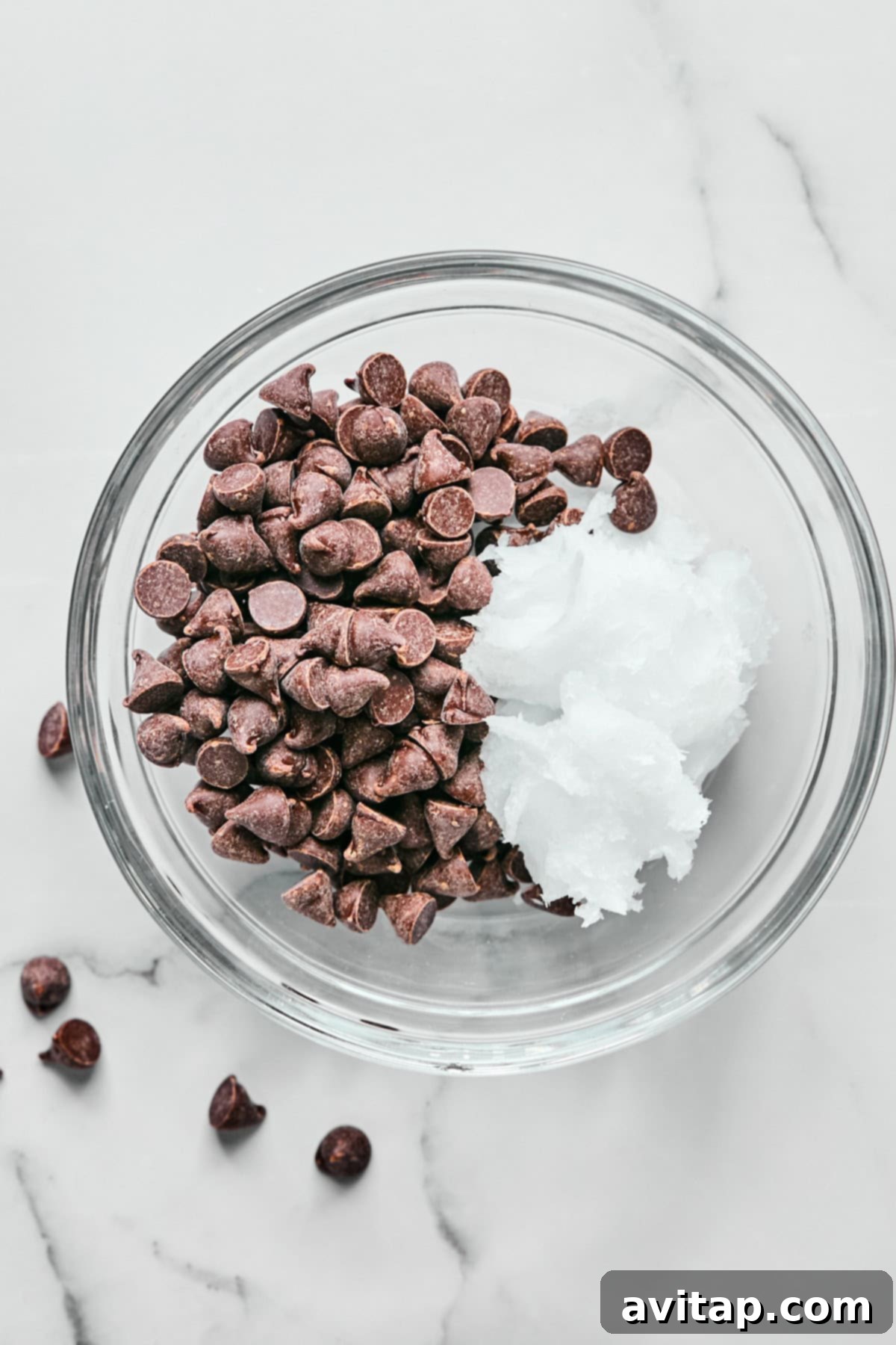A glass bowl filled with chocolate chips and a dollop of coconut oil on a marble surface.