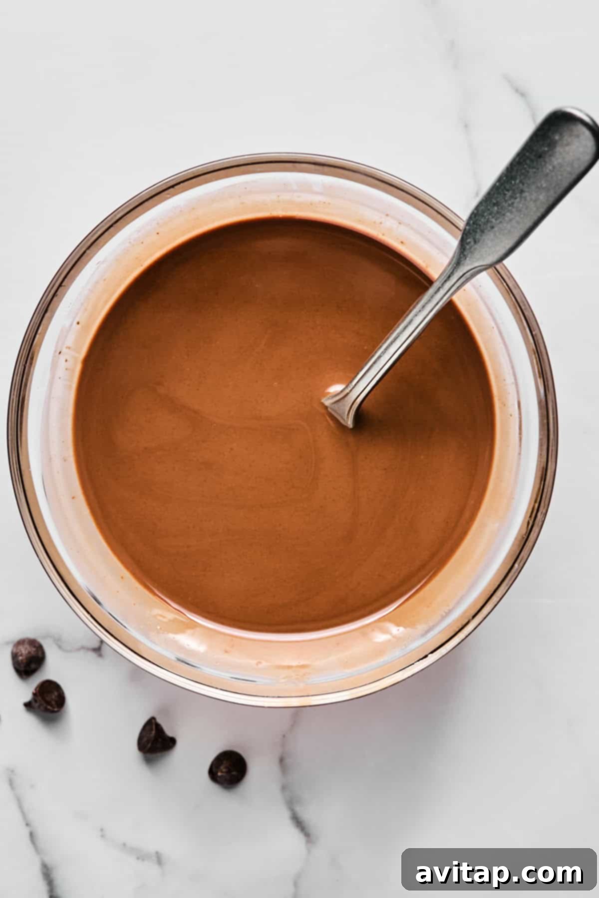 Bowl of melted chocolate with a metal spoon, placed on a white marble surface next to a few chocolate chips.