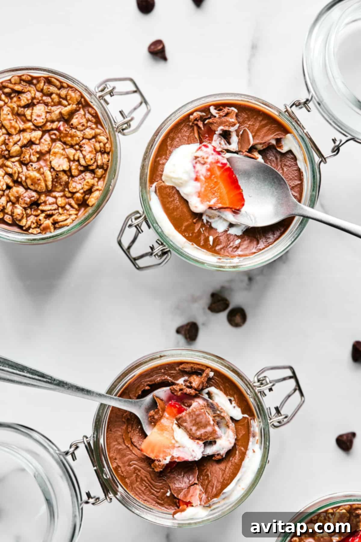 Three glass jars of chocolate mousse topped with whipped cream, strawberries, and cereal, with spoons inside. A few chocolate chips are scattered on the white surface.