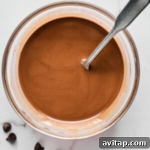 Bowl of melted chocolate with a metal spoon, placed on a white marble surface next to a few chocolate chips.
