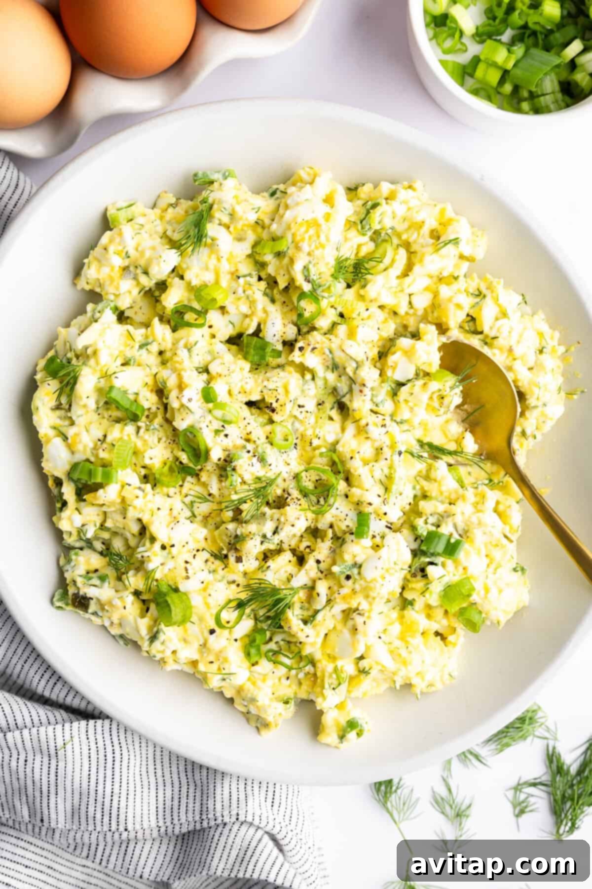 Protein-Packed Cottage Cheese Egg Salad 4 A beautifully arranged bowl of cottage cheese egg salad, elegantly garnished with freshly chopped green onions and vibrant dill. It is thoughtfully placed next to a small pile of whole boiled eggs, neatly sliced green onions, and a charming striped cloth, creating an inviting culinary scene.