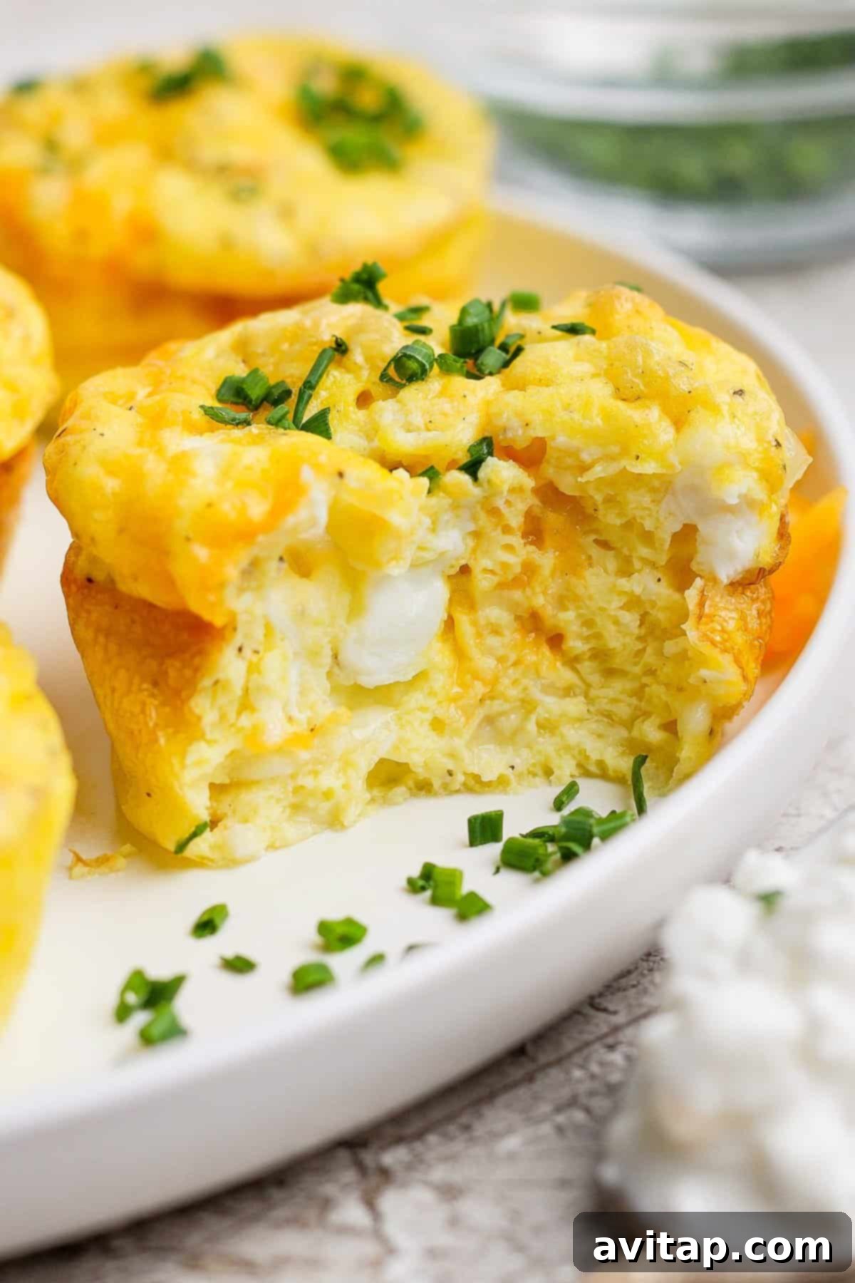 Close-up of a fluffy egg muffin topped with chopped chives, showing a bite taken out, on a white plate with blurred background.
