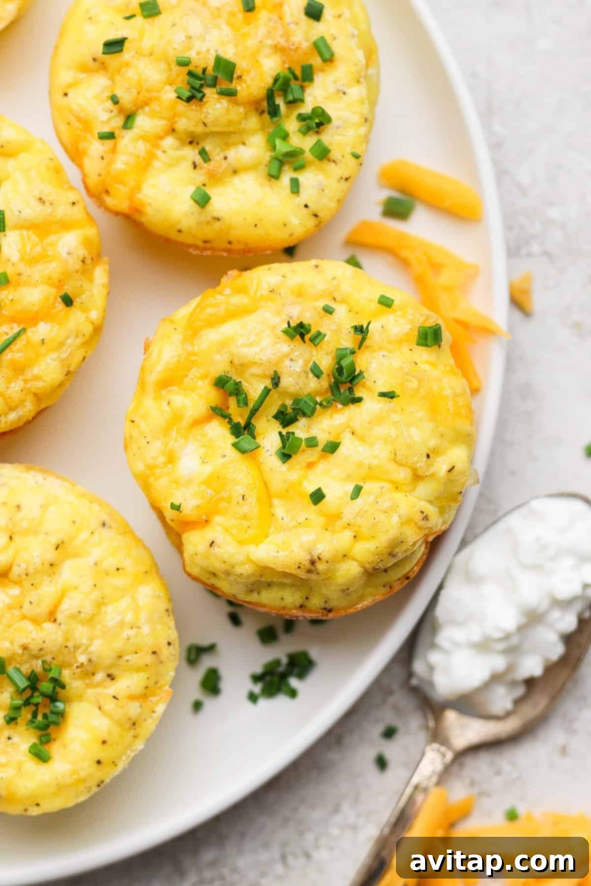 Egg muffins with cheese and chives on a white plate, accompanied by a spoon with cottage cheese and scattered cheese shreds.