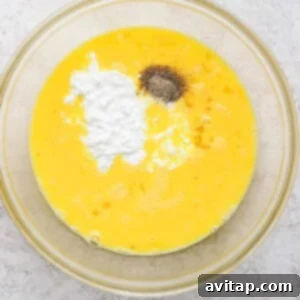A glass bowl with beaten eggs, baking powder, and black pepper on a light textured surface.