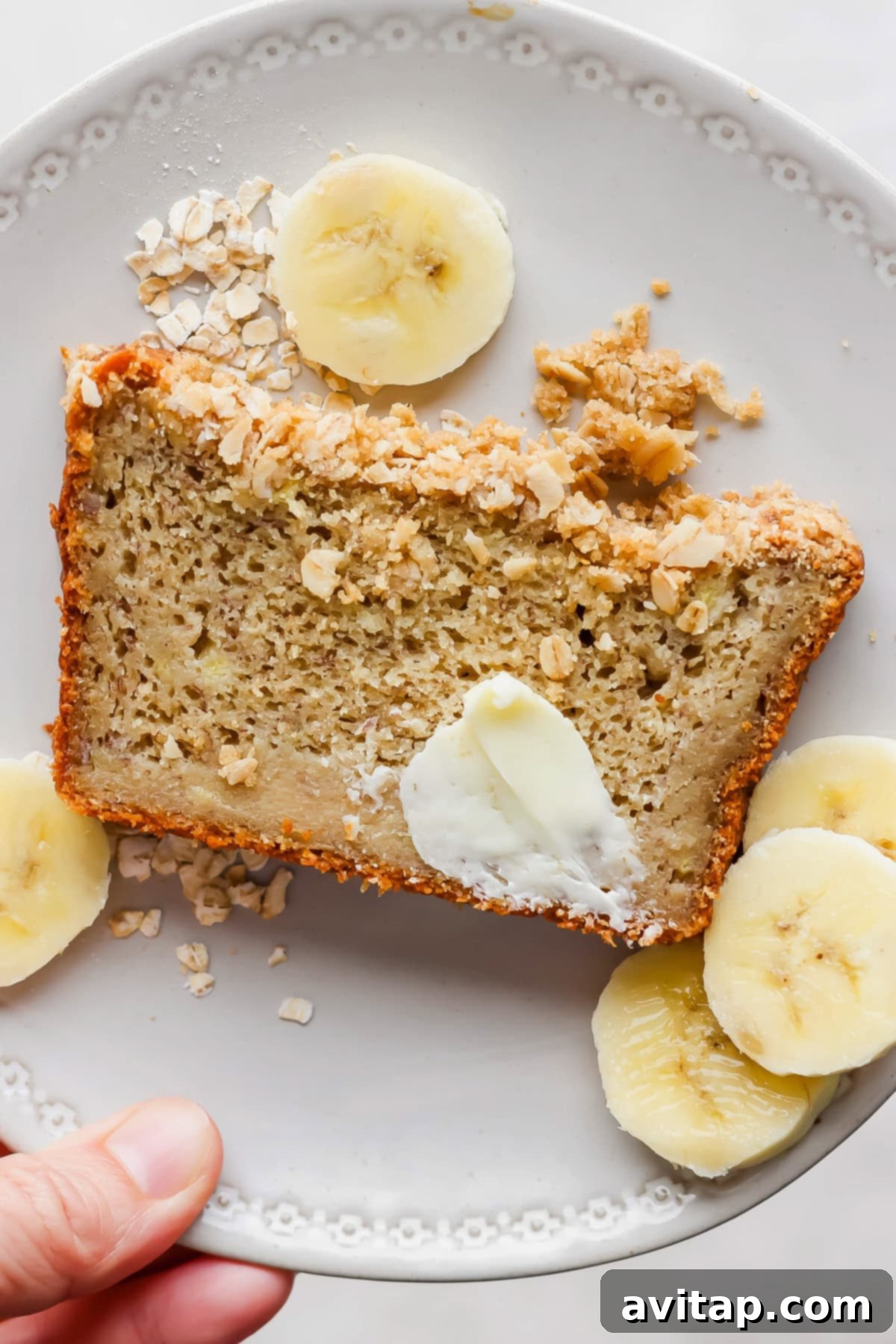 Wholesome Oat Banana Loaf 2 A delectable slice of oat flour banana bread, generously buttered and sprinkled with oats, resting on a pristine white plate, beautifully garnished with fresh banana slices.