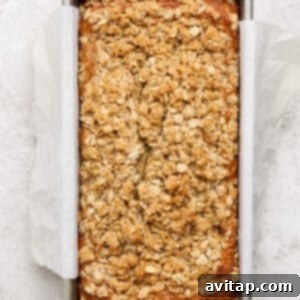 Wholesome Oat Banana Loaf 10 A freshly baked loaf of banana bread with oat crumble topping, still in its parchment-lined metal baking pan on a light textured surface, cooling gracefully.