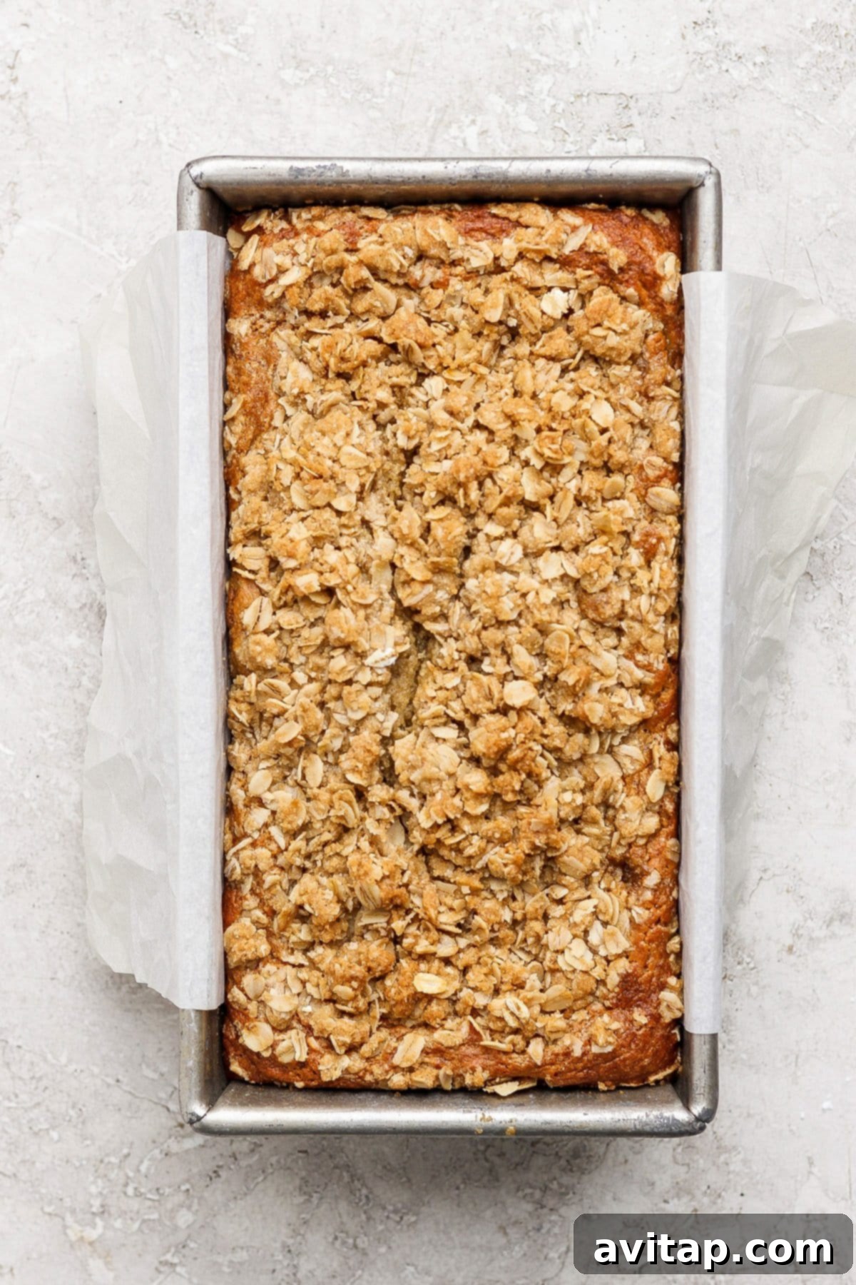 Wholesome Oat Banana Loaf 3 A freshly baked loaf of oat flour banana bread, crowned with a rustic oat crumble topping, nestled in a parchment-lined metal baking pan on a subtly textured, light-colored surface.