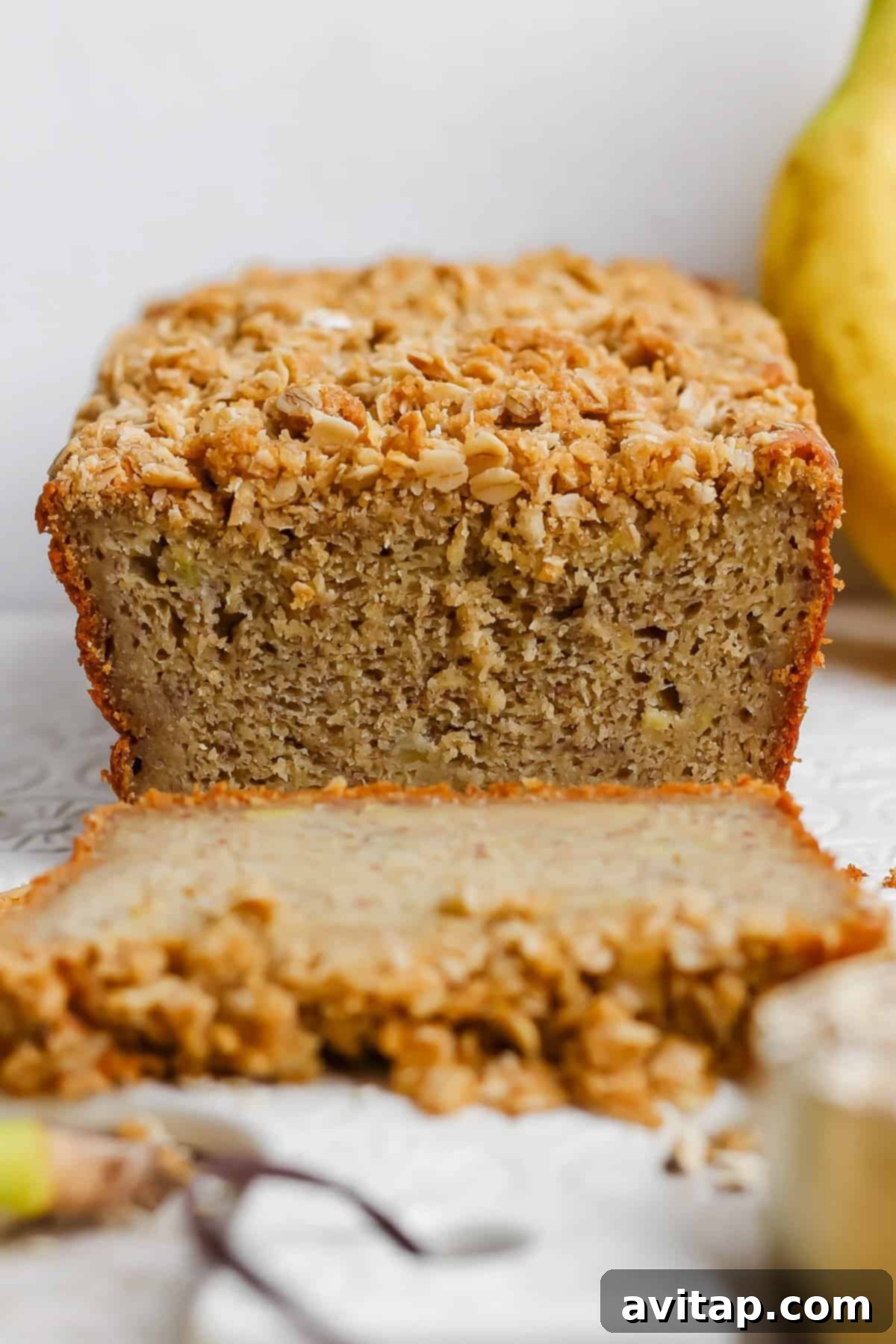 Wholesome Oat Banana Loaf 4 A beautifully sliced loaf of banana bread, generously adorned with a rustic crumbly oat and nut mixture, presented on a clean white surface, inviting a delicious indulgence.