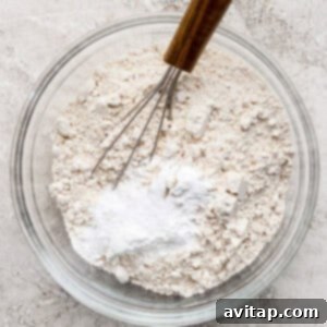 Wholesome Oat Banana Loaf 7 A glass bowl contains a mixture of flour and baking powder, with a metal whisk resting inside, placed on a textured gray surface, showcasing the dry ingredients.