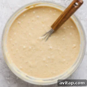 Wholesome Oat Banana Loaf 8 A glass bowl filled with thick, creamy batter is placed on a textured gray surface. A wooden-handled whisk rests inside the bowl, illustrating the wet ingredients being combined.