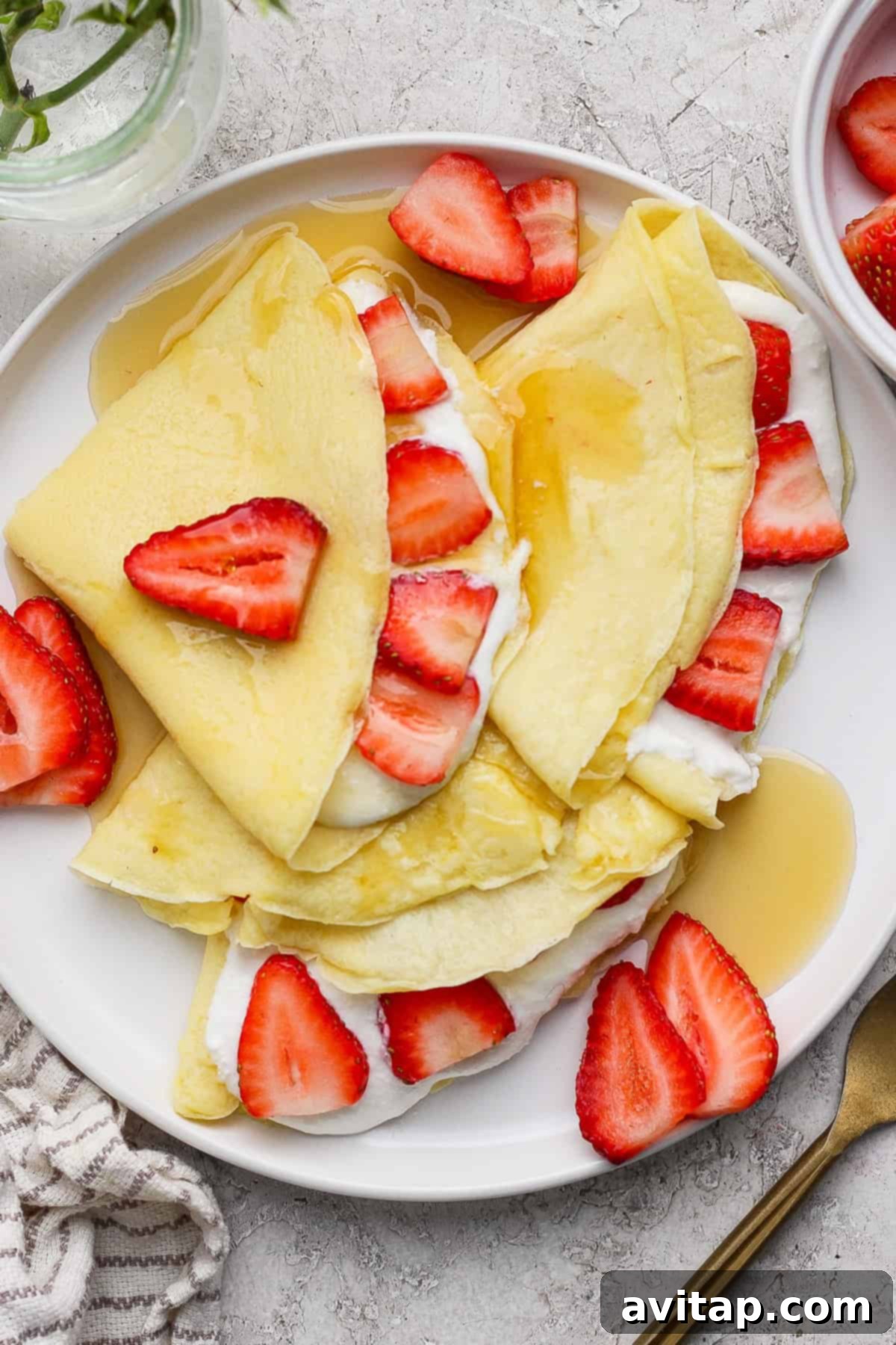 Creamy Cottage Cheese Crepes 2 Delicate cottage cheese crepes, beautifully folded and generously topped with vibrant sliced strawberries and a glistening drizzle of maple syrup, artfully arranged on a pristine white plate. An elegant fork and a charming striped napkin complete the inviting presentation.
